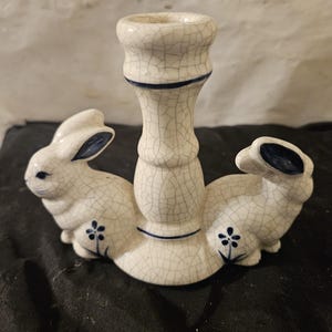 May include: A white ceramic candle holder featuring two bunny figures on either side. The candle holder has a crackle glaze finish with blue accents on the bunnies' ears and a floral design. The candle holder is a decorative item.