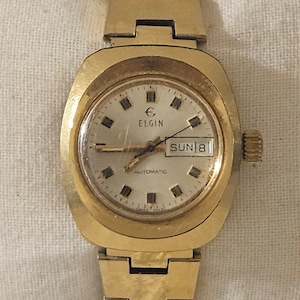 May include: A gold-toned wristwatch with a square face and a gold metal band. The watch face has black Roman numeral hour markers, a date window that reads "SUN 18", and the text "ELGIN AUTOMATIC".