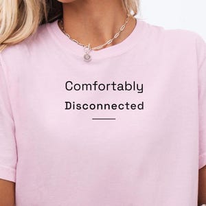 Peut inclure: T-shirt rose clair avec l'inscription "Comfortably Disconnected" en noir. Le haut a un col rond et des manches courtes. Un collier argenté avec un pendentif est visible.