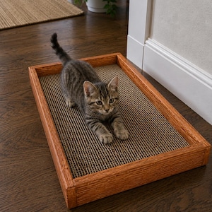 May include: A small tabby kitten with grey and brown striped fur stands on a rectangular cat scratcher. The scratcher features a textured, corrugated cardboard surface set within a sturdy, medium-toned wooden frame with a visible wood grain.