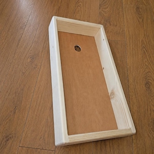 May include: A rectangular wooden tray with light-colored pine side walls and a flat, smooth brown base. The tray features a single circular hole centered near the top edge of the interior base, showcasing a simple, functional construction.