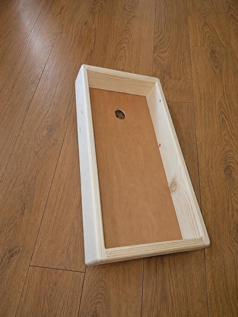 May include: A rectangular wooden tray with light-colored pine side walls and a flat, smooth brown base. The tray features a single circular hole centered near the top edge of the interior base, showcasing a simple, functional construction.
