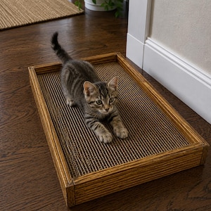 May include: A small brown tabby kitten with dark stripes stands on a rectangular cat scratcher. The scratcher features a textured, corrugated cardboard surface set within a sturdy, medium-toned wooden frame with visible wood grain patterns.
