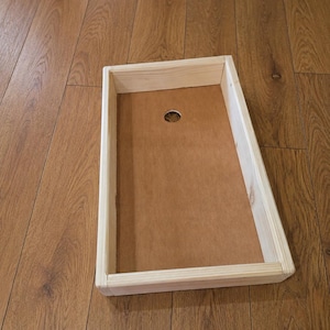 May include: A rectangular wooden cornhole board frame featuring light-colored natural wood side panels and a flat, brown, smooth surface base. A single circular hole is positioned near the top center of the board, providing a clear view of the construction.