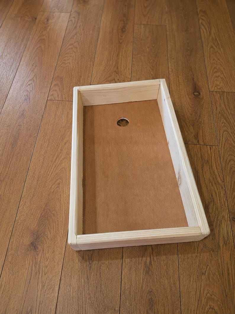 May include: A rectangular wooden cornhole board frame featuring light-colored natural wood side panels and a flat, brown, smooth surface base. A single circular hole is positioned near the top center of the board, providing a clear view of the construction.