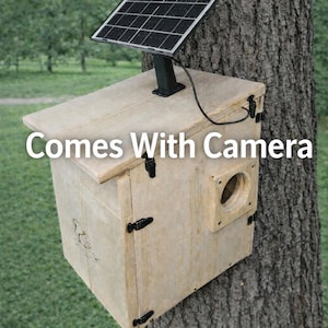 May include: A wooden birdhouse with a solar panel attached to the top. The birdhouse is mounted on a tree and has a small camera. The text "Comes With Camera" is displayed on the front of the birdhouse.