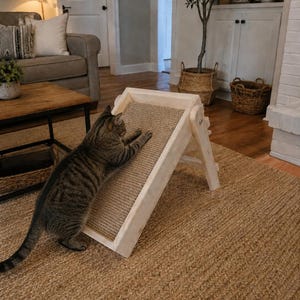 May include: A brown tabby cat stretches its front paws onto an adjustable, light-colored wooden cat scratching ramp. The ramp features a textured, tan-colored sisal scratching surface set within a sturdy, rectangular wooden frame.