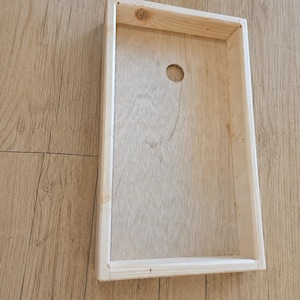 May include: A rectangular wooden cornhole board frame featuring a light natural wood grain finish. The structure includes a flat base panel with a single circular hole positioned near the top center, surrounded by four raised wooden side walls.