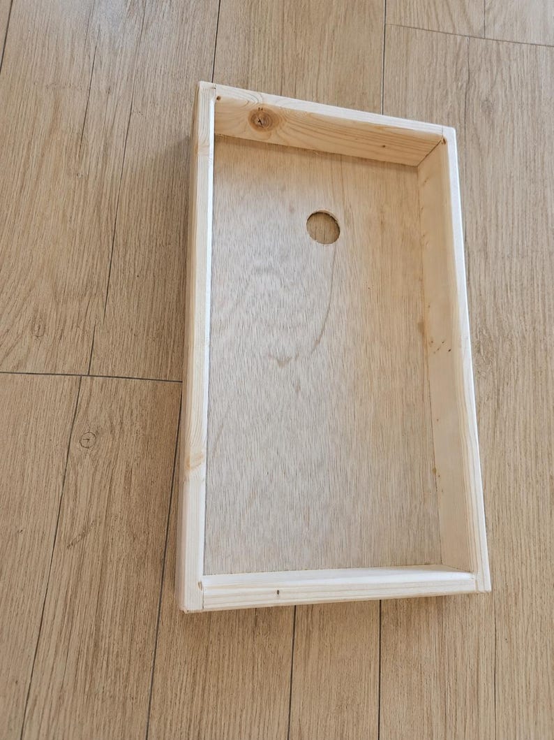 May include: A rectangular wooden cornhole board frame featuring a light natural wood grain finish. The structure includes a flat base panel with a single circular hole positioned near the top center, surrounded by four raised wooden side walls.