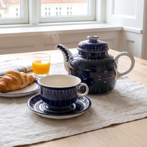 May include: A dark blue ceramic teapot, teacup, and saucer set featuring white geometric patterns. A croissant sits on a matching plate next to a glass of orange juice and a newspaper. The teapot has a lid and a handle, and the teacup rests on a saucer.