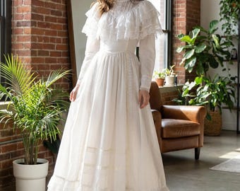 Puede incluir: Un vestido blanco largo hasta el suelo con cuello alto, mangas largas con puños y una capa superpuesta con adornos de encaje sobre el corpiño. La falda es amplia y plisada, acentuada con inserciones de encaje horizontales y un dobladillo con volantes.