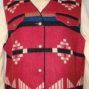 May include: A red wool vest with a Southwestern geometric pattern. The vest features black, white, and tan accents, two pockets, and decorative silver buttons. The vest is worn over a yellow collared shirt.