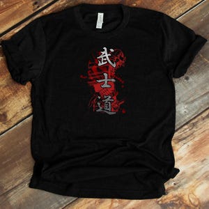 Puede incluir: Camiseta negra con un diseño de inspiración japonesa. El gráfico presenta caracteres kanji en blanco y gris sobre un fondo rojo. La camiseta está hecha de un tejido suave y tiene un cuello redondo clásico.