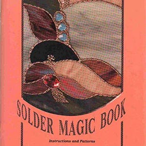 May include: A softcover book titled Solder Magic Book: Instructions and Patterns by Kay Bain Weiner. The cover features a coral-colored background with a central circular graphic displaying a stained glass design in shades of purple, blue, and tan.