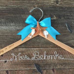 May include: A wooden hanger with a blue bow and a starfish, seashells, and the text "July 29, 2017". The hanger is decorated with the text "Mrs. Behmke" in silver wire.