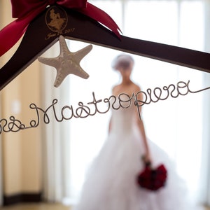 Puede incluir: Una percha de madera con un lazo rojo y una decoraci&oacute;n de estrella de mar. La percha tiene las palabras "Mrs. Mastropiero" escritas en alambre.
