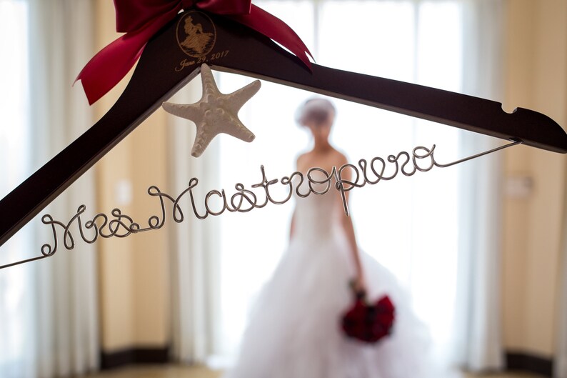 Puede incluir: Una percha de madera con un lazo rojo y una decoraci&oacute;n de estrella de mar. La percha tiene las palabras "Mrs. Mastropiero" escritas en alambre.
