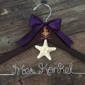 Puede incluir: Una percha de madera marr&oacute;n con un lazo de cinta morado y una estrella de mar. La percha est&aacute; personalizada con el texto "Mrs. Konkel" y una silueta de Ariel de La Sirenita. La fecha "June 11, 2016" tambi&eacute;n est&aacute; grabada en la percha.