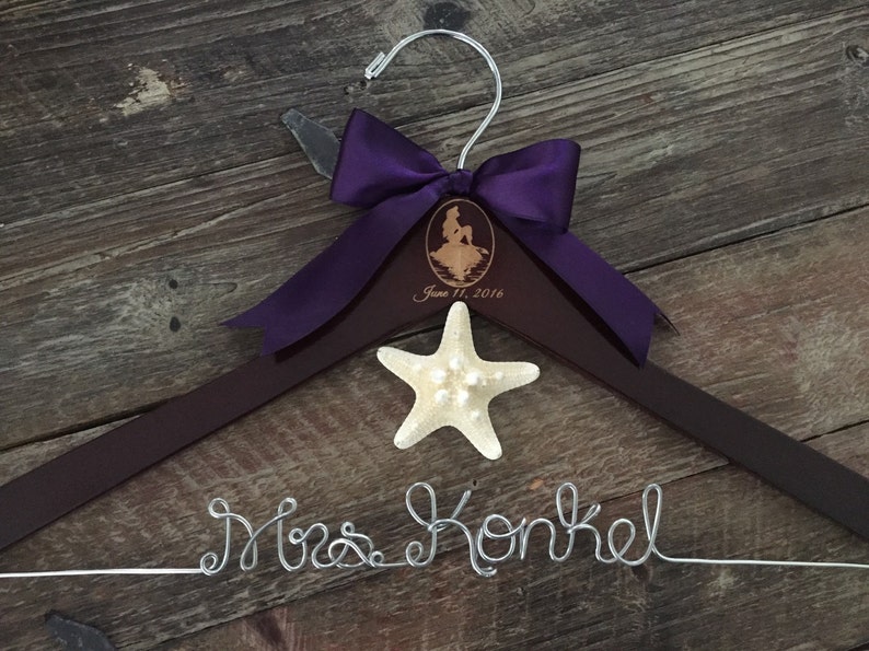 Puede incluir: Una percha de madera marr&oacute;n con un lazo de cinta morado y una estrella de mar. La percha est&aacute; personalizada con el texto "Mrs. Konkel" y una silueta de Ariel de La Sirenita. La fecha "June 11, 2016" tambi&eacute;n est&aacute; grabada en la percha.