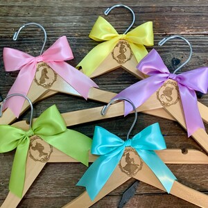 May include: Five wooden hangers with different colored satin bows. Each hanger has a laser-engraved design of a Disney princess silhouette.