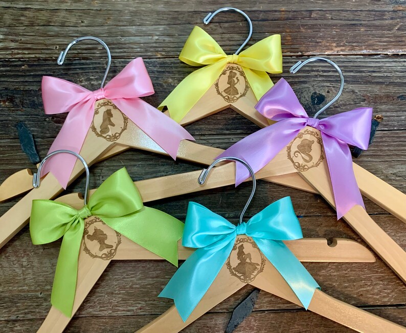 May include: Five wooden hangers with different colored satin bows. Each hanger has a laser-engraved design of a Disney princess silhouette.