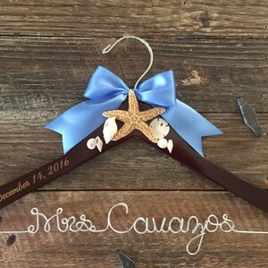May include: A brown wooden hanger with a blue bow and a starfish design. The hanger is engraved with the text "December 14, 2016" and "Mrs. Cavanos".