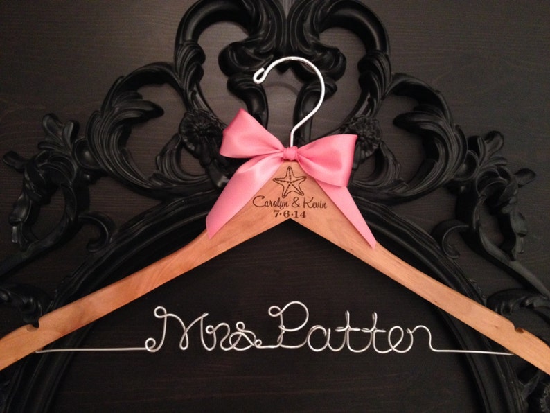May include: Wooden hanger with a pink bow and the text "Carolyn & Kevin 7-0-14" engraved on the top. The hanger has a silver wire inscription that reads "Mrs. Patten".