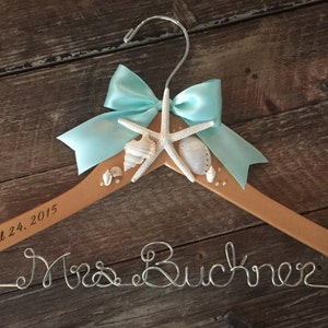 May include: A wooden hanger decorated with a blue bow, seashells, and a starfish. The hanger is engraved with the text "April 24, 2015" and "Mrs. Buckner".