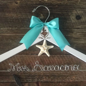 Puede incluir: Percha de madera blanca con un lazo turquesa, una estrella de mar y el texto "Mrs. Saracino" en alambre plateado. La percha tambi&eacute;n tiene una peque&ntilde;a etiqueta redonda con una ilustraci&oacute;n de sirena y el texto "May 28, 2017".