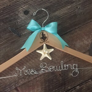 Puede incluir: Una percha de madera con un lazo turquesa, una estrella de mar y el texto "Mrs. Bowling" en alambre plateado. La percha est&aacute; personalizada con una silueta de sirena y la fecha "April 1, 2016".