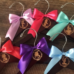 May include: Six wooden hangers with satin bows in various colors, each featuring a gold silhouette of a Disney princess. The hangers are ideal for hanging children's clothing.
