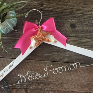 May include: White wooden hanger with a pink bow and a starfish decoration. The hanger is engraved with the text "May 17, 2017" and "Mrs. Forman".