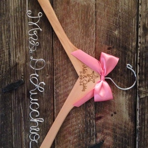 May include: A wooden hanger with a pink bow and a starfish design. The hanger is engraved with the text "Nate & Ashley 3-22-14". The hanger also has a wire inscription that reads "Mrs. DeLucchio".