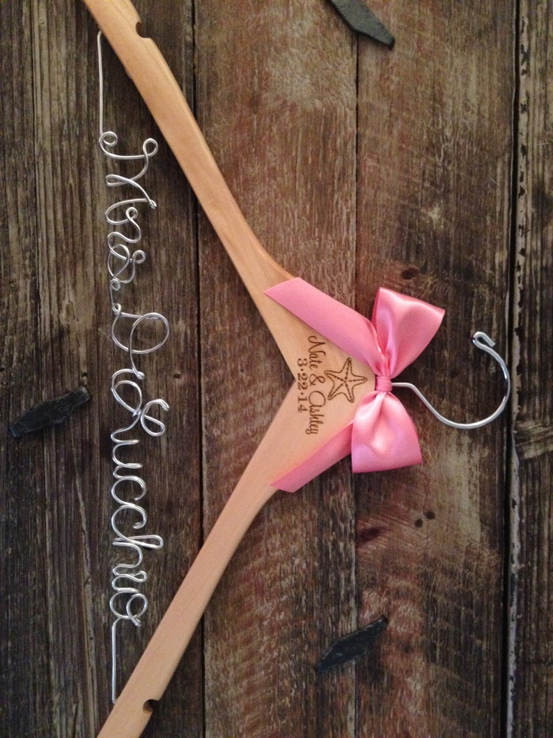 May include: A wooden hanger with a pink bow and a starfish design. The hanger is engraved with the text "Nate & Ashley 3-22-14". The hanger also has a wire inscription that reads "Mrs. DeLucchio".