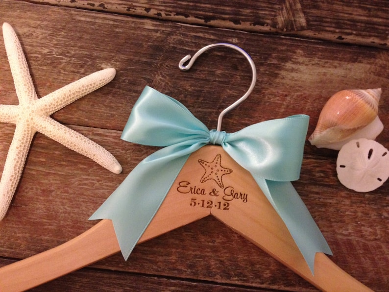 May include: A wooden hanger with a light blue satin bow and a starfish design. The hanger is personalized with the text "Erica & Gary 5-12-12".