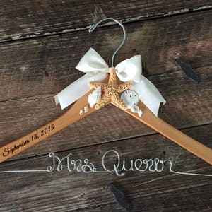 May include: A wooden hanger with a white satin bow and a starfish with seashells. The hanger is engraved with "September 18, 2015" and "Mrs. Queen" in wire.