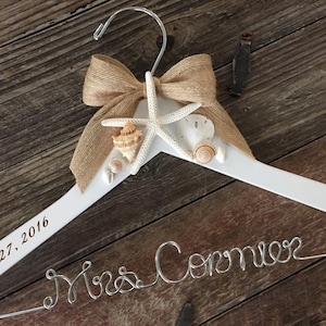 May include: White wooden hanger with a burlap bow and seashells. The hanger is engraved with the text "May 27, 2016" and "Mrs. Cormier" in silver wire.