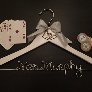 May include: White wooden hanger with a silver wire inscription that reads "Mrs. Murphy". The hanger has a silver bow and a wooden sign that reads "Welcome to Las Vegas". There are playing cards and poker chips on the surface.