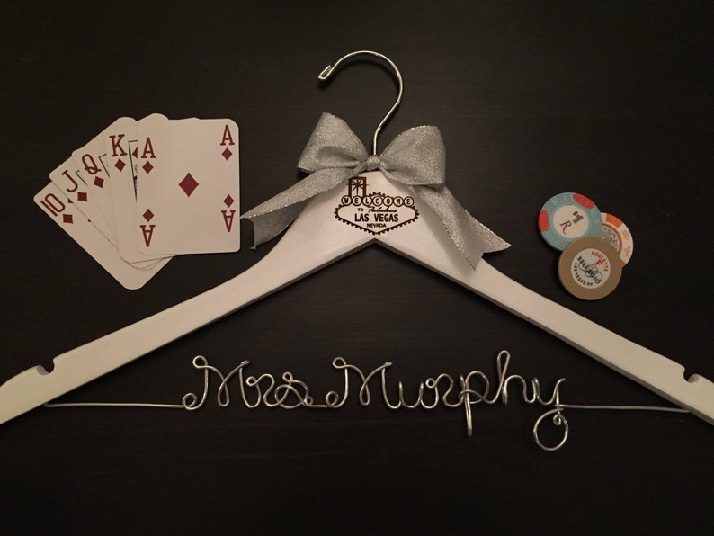 May include: White wooden hanger with a silver wire inscription that reads "Mrs. Murphy". The hanger has a silver bow and a wooden sign that reads "Welcome to Las Vegas". There are playing cards and poker chips on the surface.