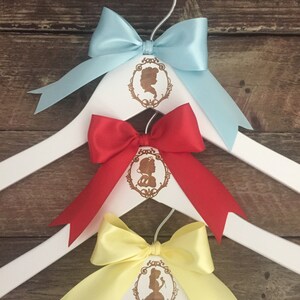 May include: Three white wooden hangers with a princess silhouette design. Each hanger has a different colored satin bow: light blue, red, and yellow.