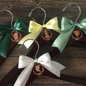 May include: Four wooden hangers with satin ribbon bows in green, yellow, white, and sage green. Each hanger has a laser-engraved design of a woman's silhouette in a frame.