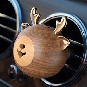 May include: A wooden car air freshener in the shape of a reindeer head, featuring gold-coloured antlers and accents. The round air freshener has a carved face and is set against a car's air vent.