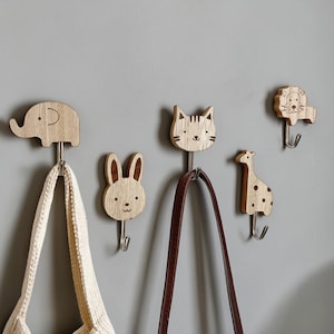 Può includere: Cinque ganci da parete in legno a forma di animali. I ganci hanno la forma di un elefante, un coniglio, un gatto, una giraffa e un leone. Ogni gancio ha un gancio in metallo argentato. Una borsa bianca e una borsa marrone sono appese ai ganci.