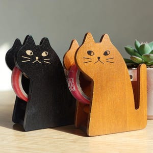 May include: Two cat-shaped tape dispensers. One is black, the other is a light brown color. Each dispenser holds a roll of tape. The cat faces have simple drawn features. A small succulent plant is in the background.