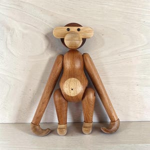 Könnte beinhalten: Holz-Affenfigur mit beweglichen Gliedmaßen. Der Affe hat einen dunkelbraunen Kopf und Körper mit hellbraunen Armen, Beinen und Gesicht. Die Arme des Affen sind ausgestreckt, und er hat einen runden Bauch mit einem kleinen Loch. Die Füße des Affen sind gebogen.