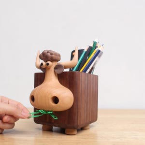 Op de afbeelding: Een houten bureau-organizer in de vorm van een stier, met een lichtbruine kop en een donkerbruin lichaam. De neus van de stier houdt groene paperclips vast. De organizer bevat potloden en pennen. De stier heeft een grillig ontwerp.