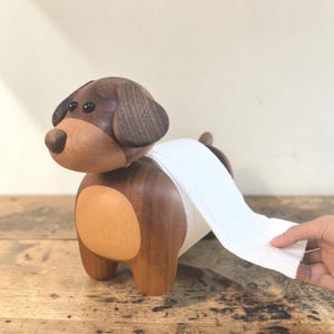 May include: A wooden dog-shaped toilet roll holder. The dog is crafted from dark and light brown wood, featuring a light brown belly and dark brown head and ears. White toilet roll is dispensed from the dog's back.