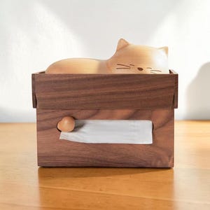 Op de afbeelding: Houten tissuebox met een kattenontwerp. De rechthoekige doos heeft een donkerbruine afwerking en een kleine houten knop aan de zijkant. Witte tissues zijn zichtbaar. De kat is lichtbruin.
