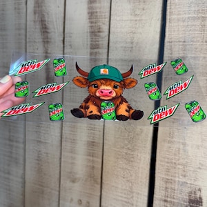 May include: A clear sticker featuring a cartoon Highland cow wearing a green cap and holding a green can of Mtn Dew. The sticker also includes multiple Mtn Dew logos and cans in a repeating pattern.