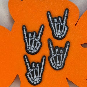 May include: Three individual skeleton hand emblems arranged on a bright orange leaf-shaped base. Each emblem features a white skeletal hand making the rock and roll sign with black outlines and detailing, set against a solid orange background.